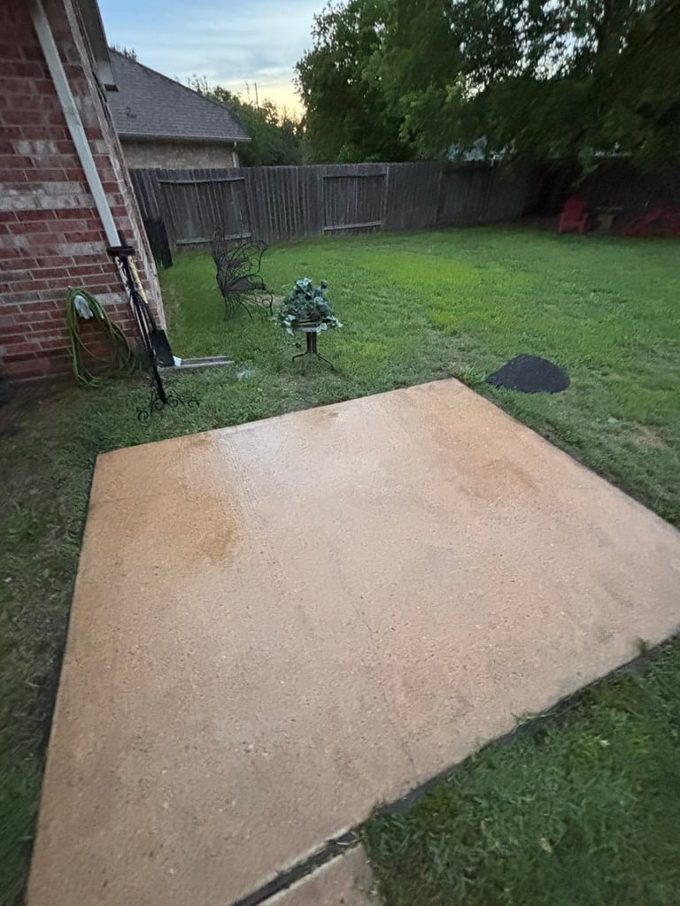 Clean concrete patio in green backyard with wooden fence, brick house corner, and garden tools visible