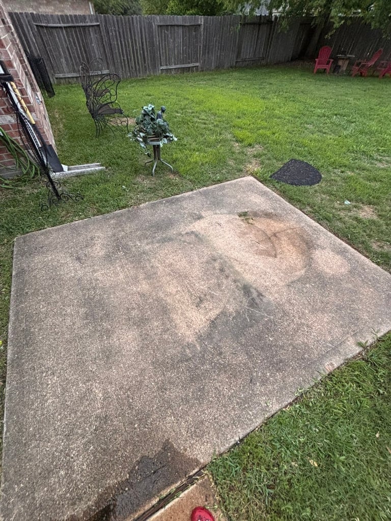 Weathered concrete patio in backyard with grass, wooden fence, metal chair, and potted plant in background